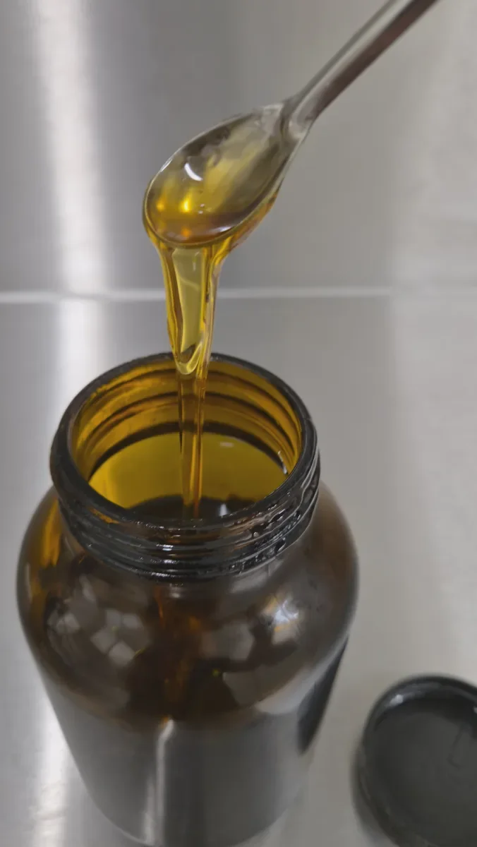 Golden Full Spectrum hemp distillate flowing from a spoon into an amber glass jar on a stainless steel worktop