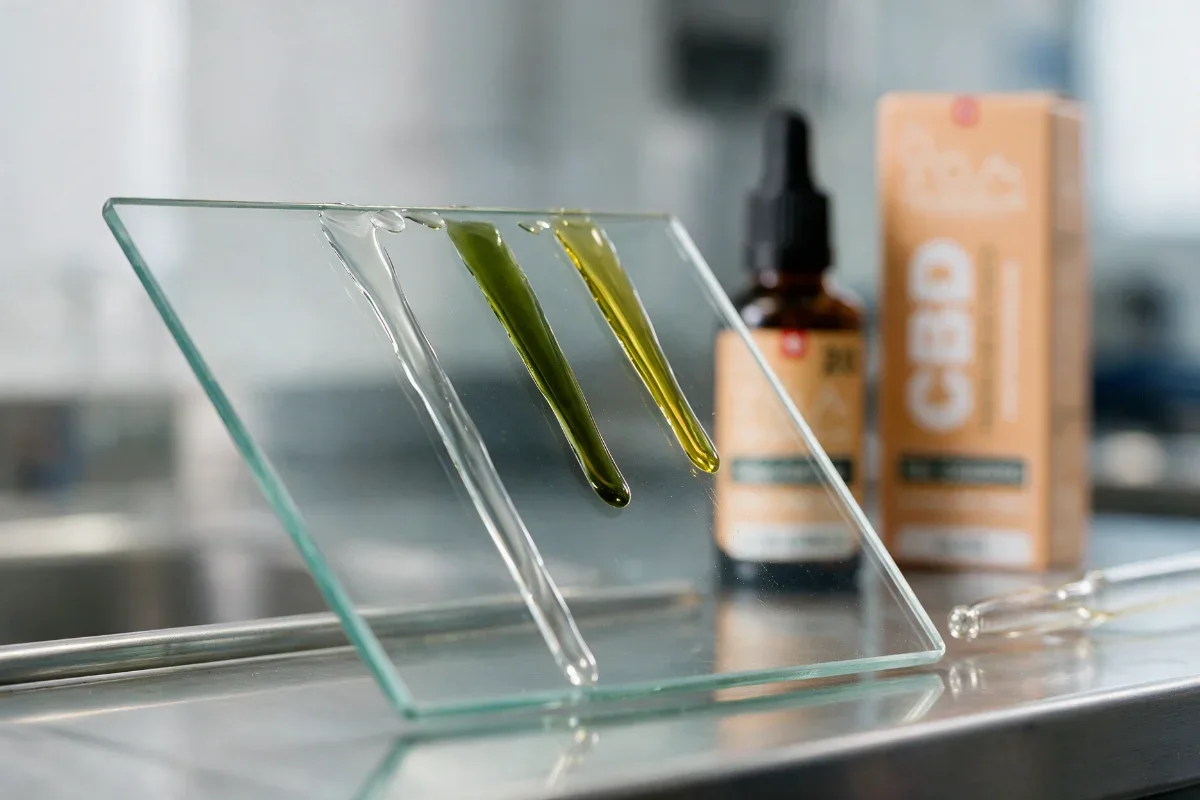 Drei Öltropfen auf einer geneigten Glasplatte: Die Spur des farblosen MCT ist am längsten, die des grünen Hanföls mittellang, die des goldenen Olivenöls am kürzesten