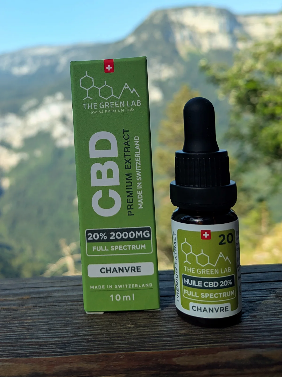 Pipettenfläschchen mit CBD-Hanföl 20 % Full Spectrum und grüner Schachtel auf einem Holzzaun vor Bergkulisse