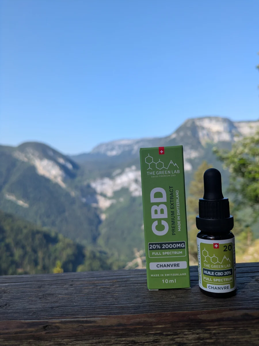 Pipettenfläschchen mit CBD-Hanföl 20 % Full Spectrum und grüner Schachtel auf einem Holzzaun vor Bergkulisse