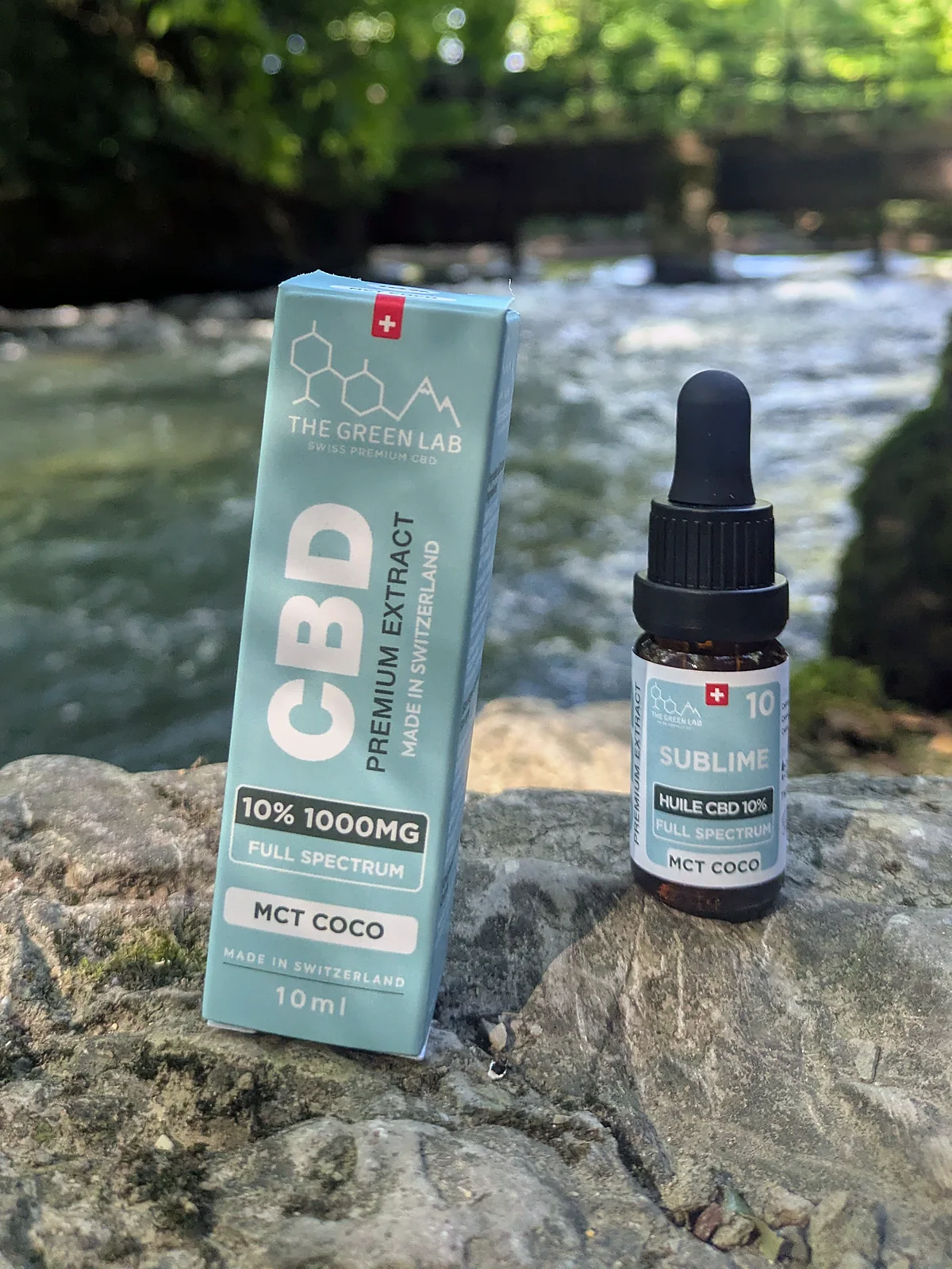 Tropfflasche mit CBD-Öl Sublime 10 % auf MCT-Kokosöl und hellblauer Schachtel, auf einem Felsen am Ufer eines Flusses