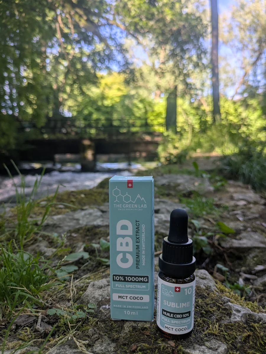 Tropfflasche mit CBD-Öl Sublime 10 % auf MCT-Kokosöl und hellblauer Schachtel, auf einem Felsen am Ufer eines Flusses