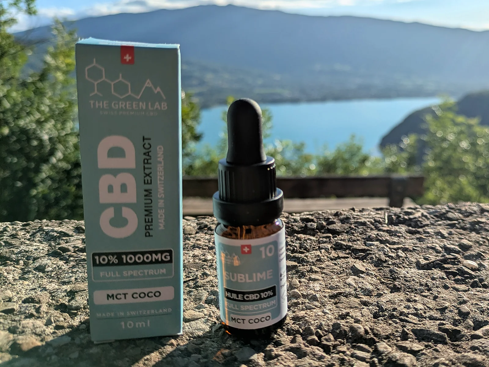 Faltschachtel und Flasche CBD-Öl Sublime 10 % MCT-Kokos auf einer Mauer, See und Berge im Hintergrund
