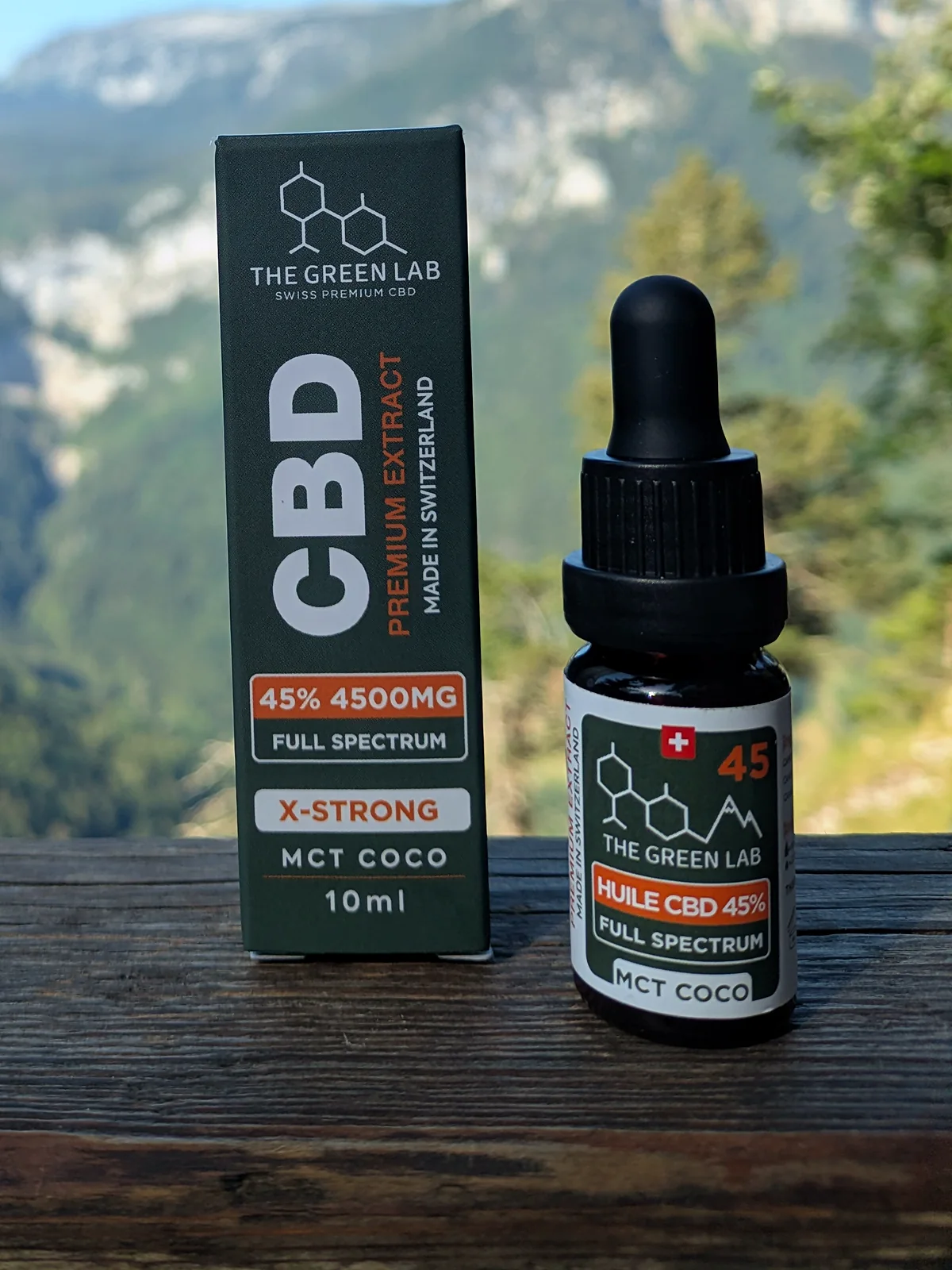 Pipettenfläschchen mit CBD-Öl X-Strong 45 % Full Spectrum und anthrazit-oranger Schachtel vor einer Berglandschaft