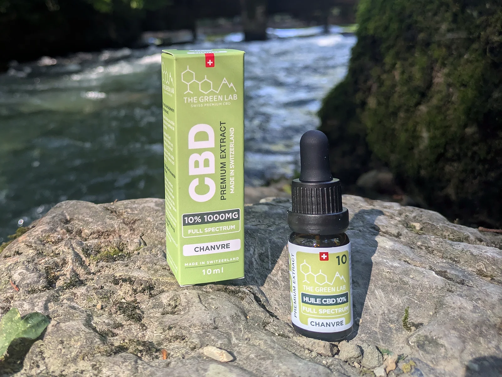 Flacon compte-gouttes d'huile CBD Chanvre 10 % et son étui vert clair sur un rocher, au bord d'une rivière