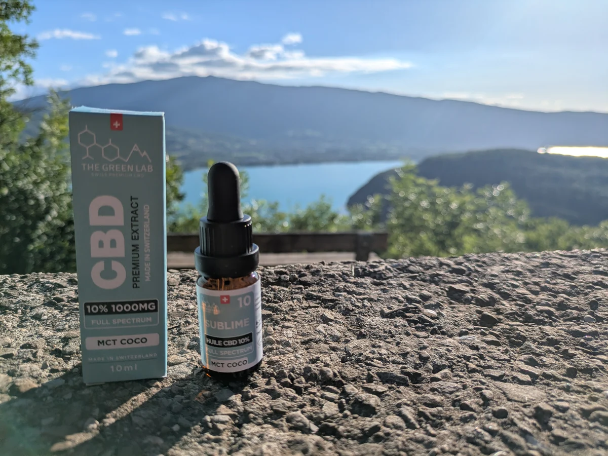 Étui et flacon d'huile CBD Sublime 10 % MCT coco posés sur un muret, lac et montagnes en arrière-plan